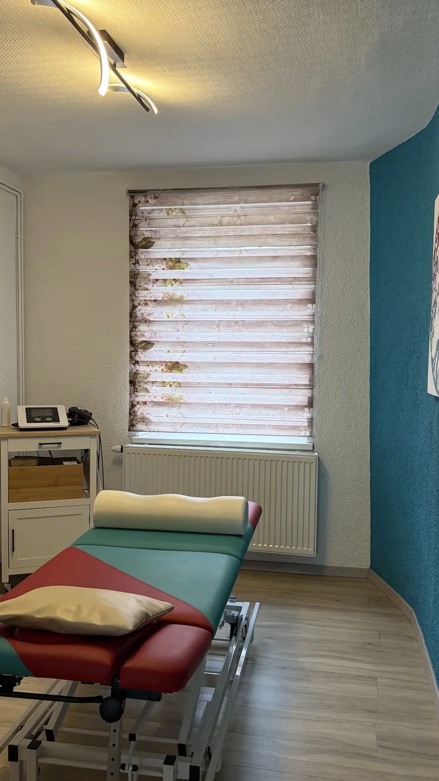 Behandlungsraum der Praxis Gurski – Blick auf Liege und florales Rollo